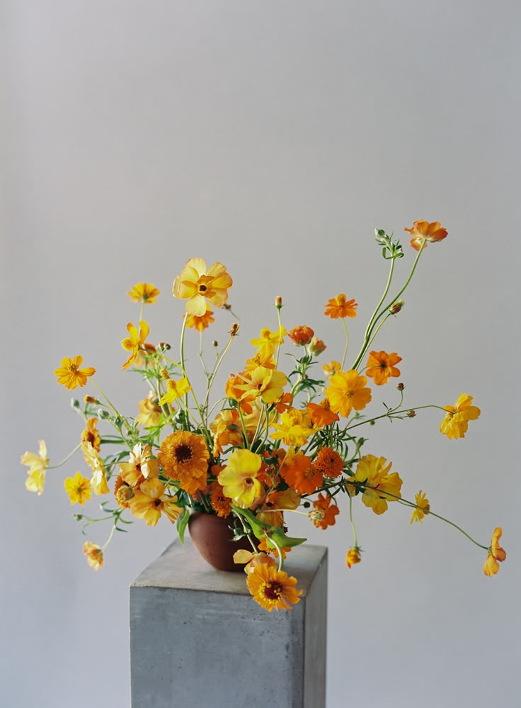 a vase filled with yellow and orange flowers on top of a cement block in front of a white wall