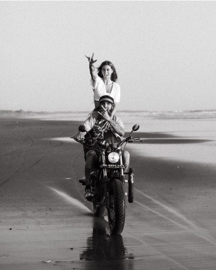 a man and woman riding on the back of a motorcycle down a wet beach road