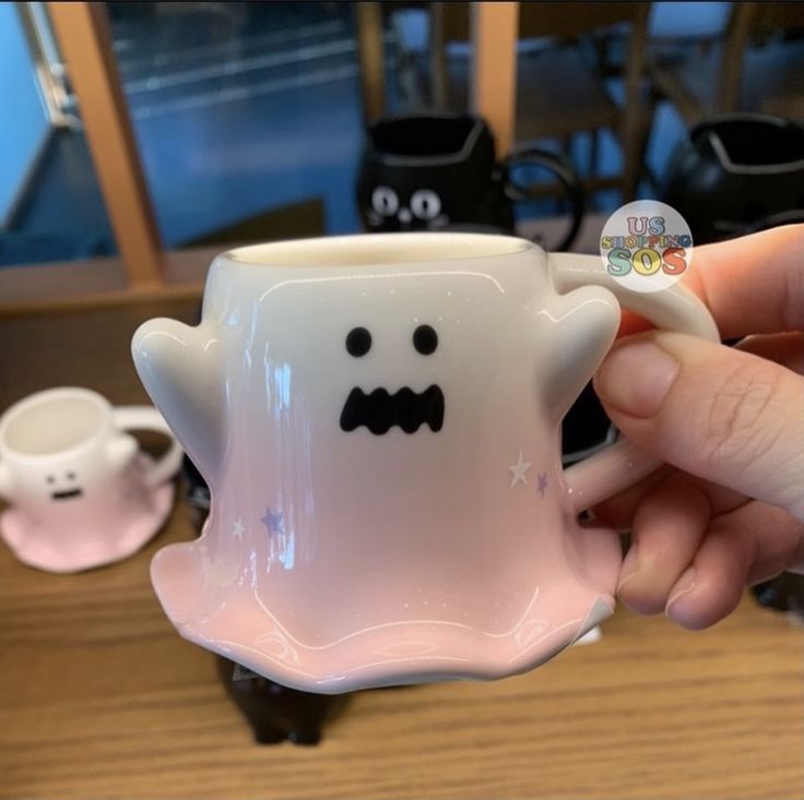 a hand holding a coffee cup with a ghost face on it and two other cups in the background