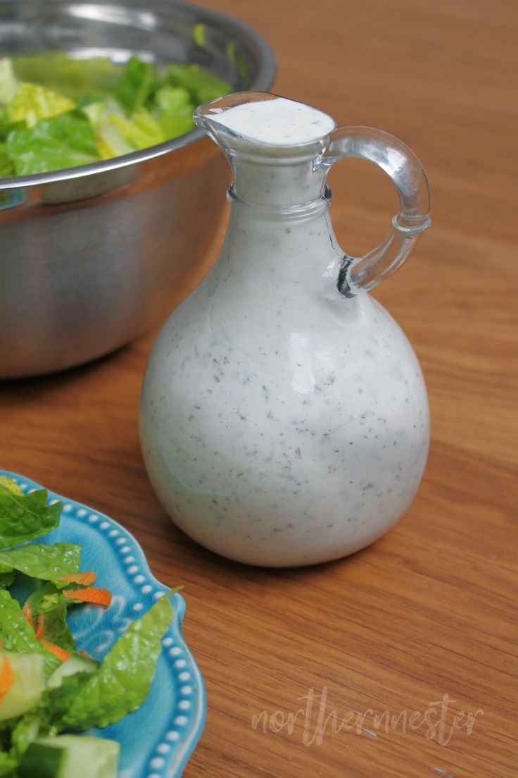 a salad in a bowl next to a pitcher of dressing on a wooden table top