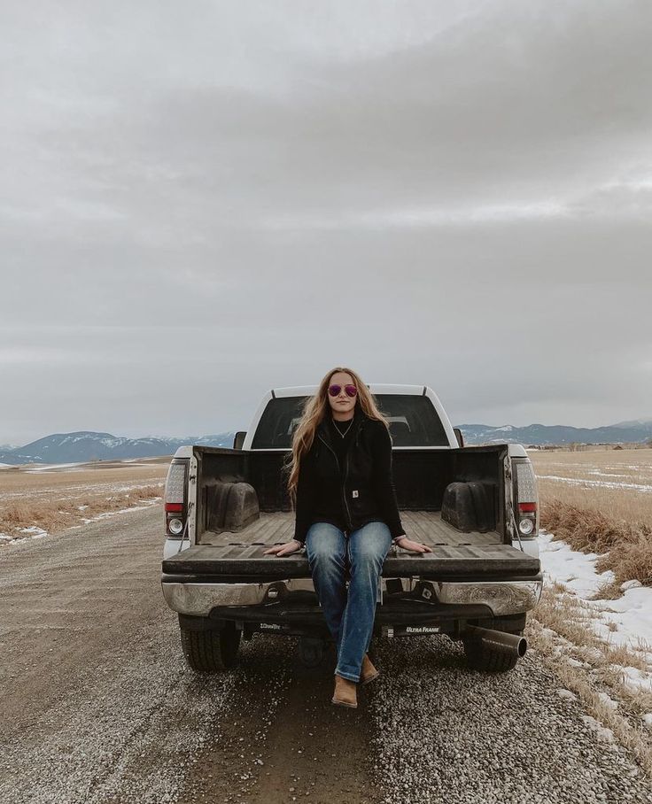 a woman sitting in the back of a truck on top of a dirt road next to a field