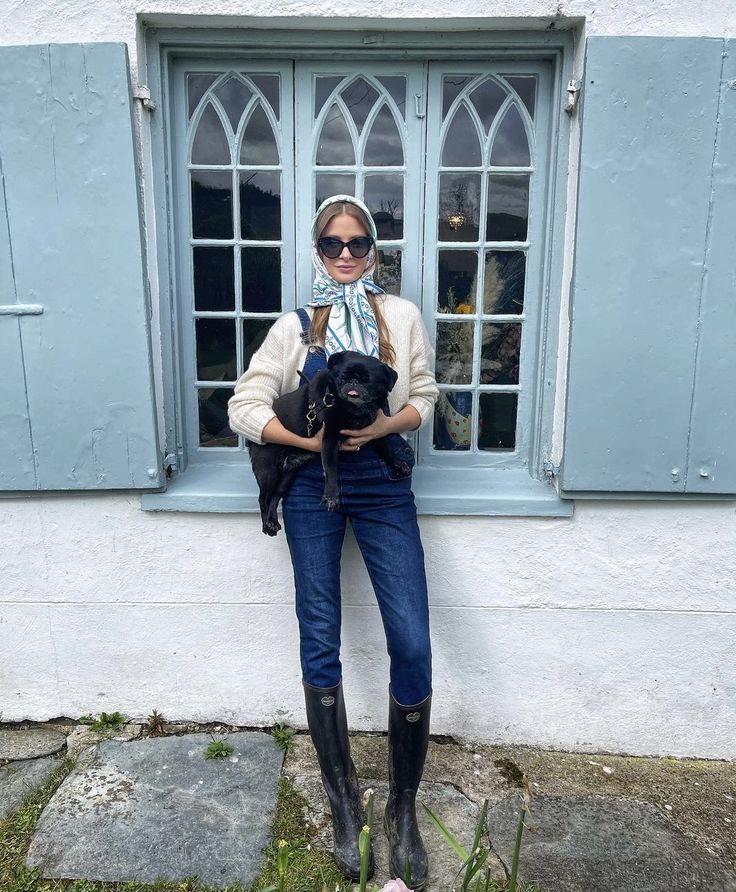 a woman standing in front of a blue window holding a black dog wearing a scarf