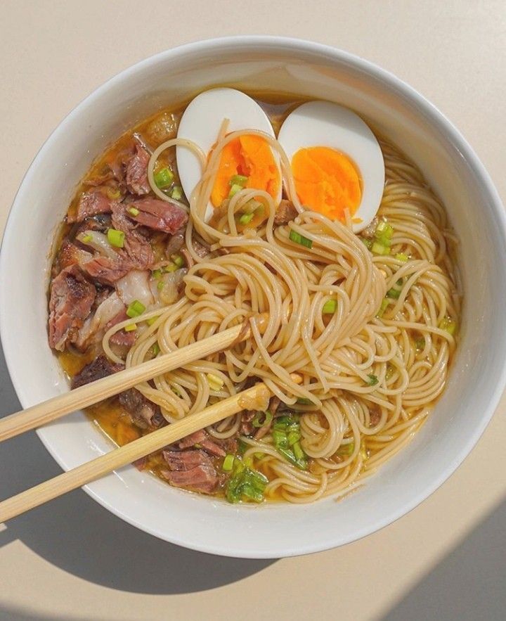 a bowl filled with noodles, meat and eggs on top of a wooden table next to chopsticks