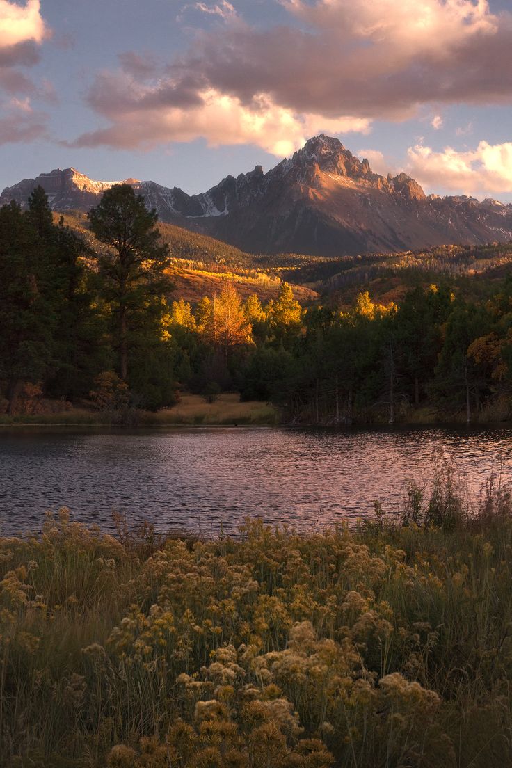 a lake surrounded by mountains and trees under a cloudy sky with clouds in the background