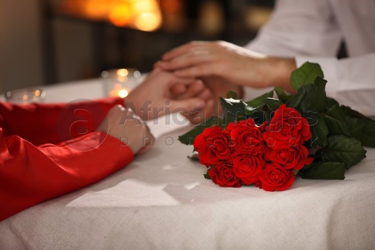 two people holding hands over a table with red roses on it and candles in the background