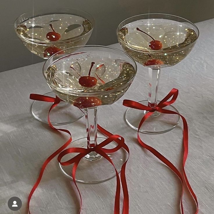 three wine glasses with cherries in them and red ribbon on the rim, sitting on a table