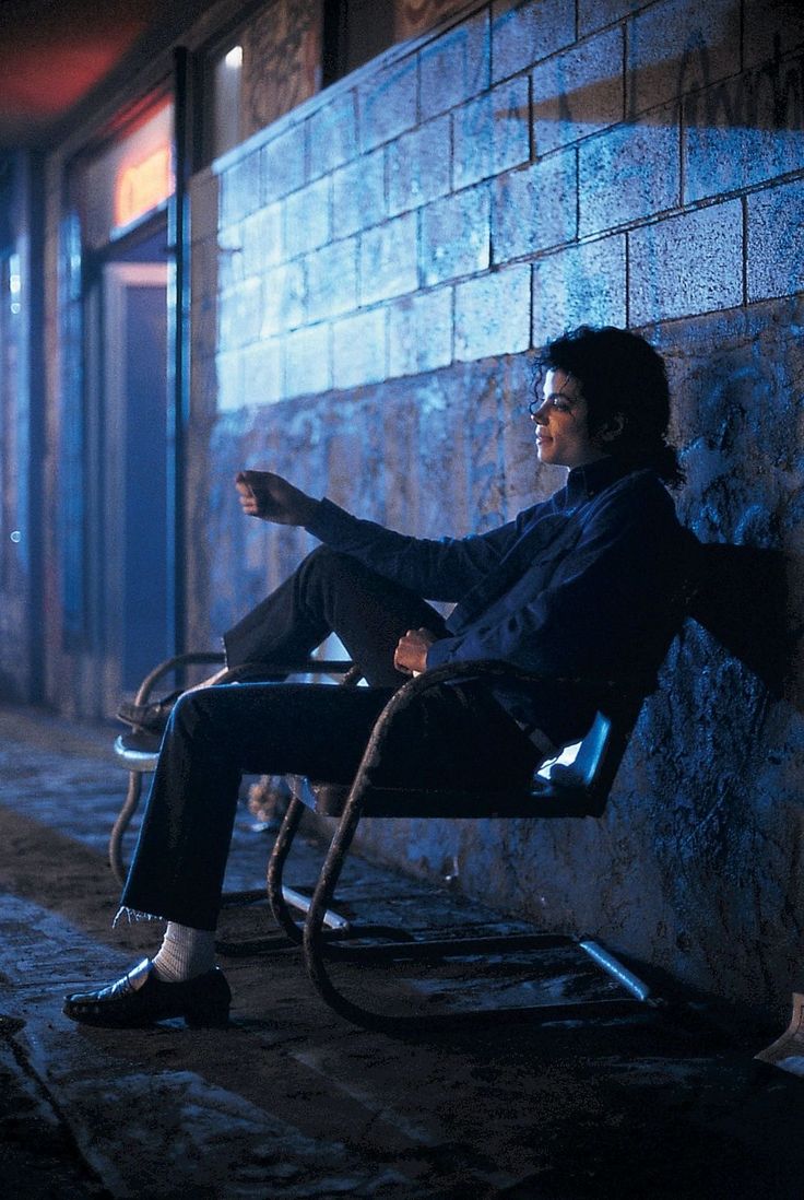 a man sitting on a bench in front of a building at night with his hand up