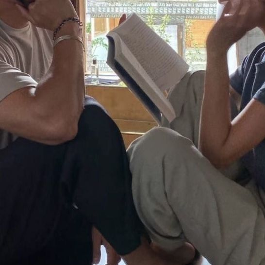 a man sitting on the floor reading a book while holding a cell phone to his ear