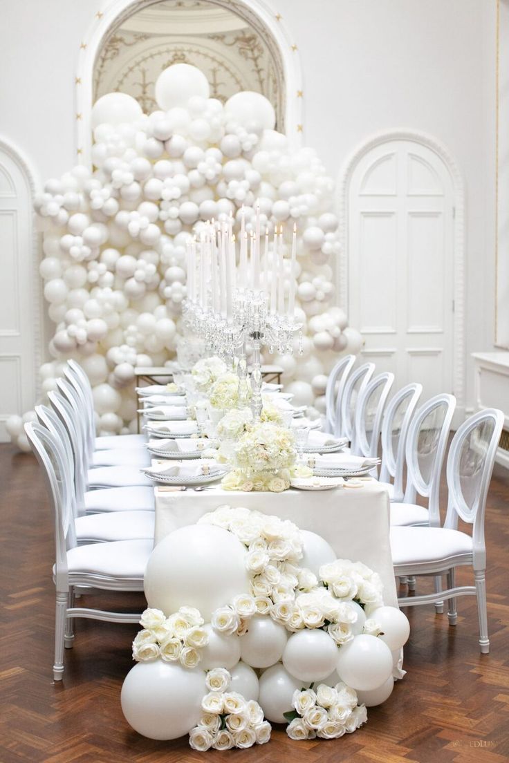 a long table with white balloons and flowers on it is set for an elegant dinner