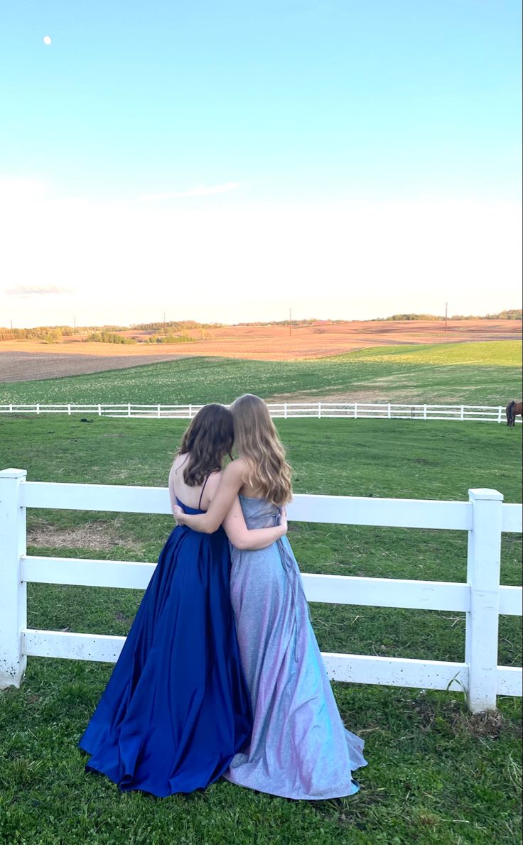 two women in long dresses standing next to a white fence looking out at the field