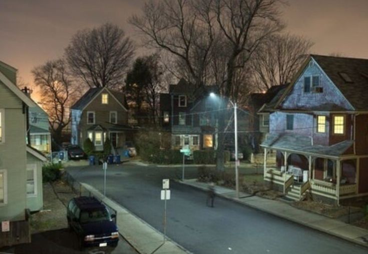 an empty street at night with some houses in the background and one car parked on the side