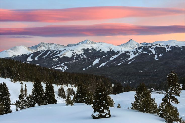 the mountains are covered in snow as the sun is setting over them and pink clouds loom overhead