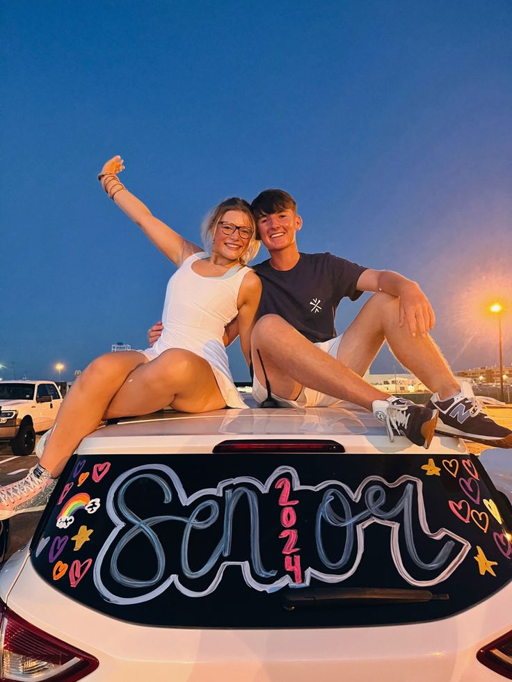 a man and woman sitting on the hood of a car with their arms in the air