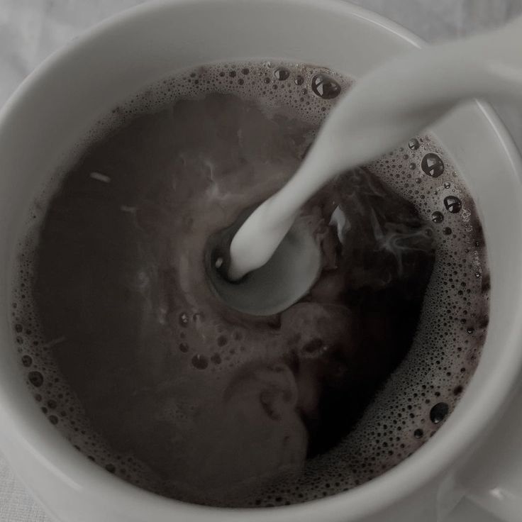 milk being poured into a coffee cup filled with brown liquid on top of a white table cloth