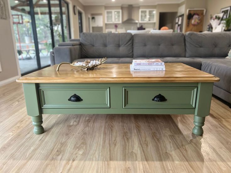 a green coffee table with two drawers in front of a gray couch and large windows