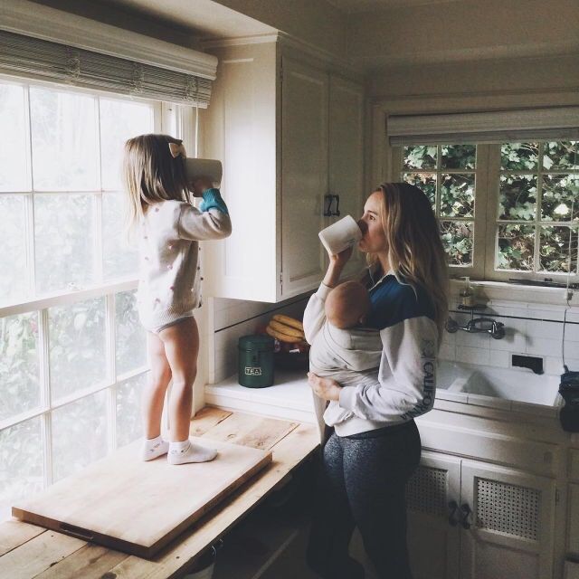 two women and a child are standing on a kitchen counter drinking from cups in front of the window