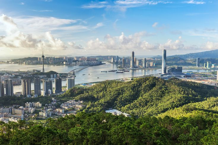 an aerial view of a city and the ocean with mountains in the foreground, surrounded by greenery