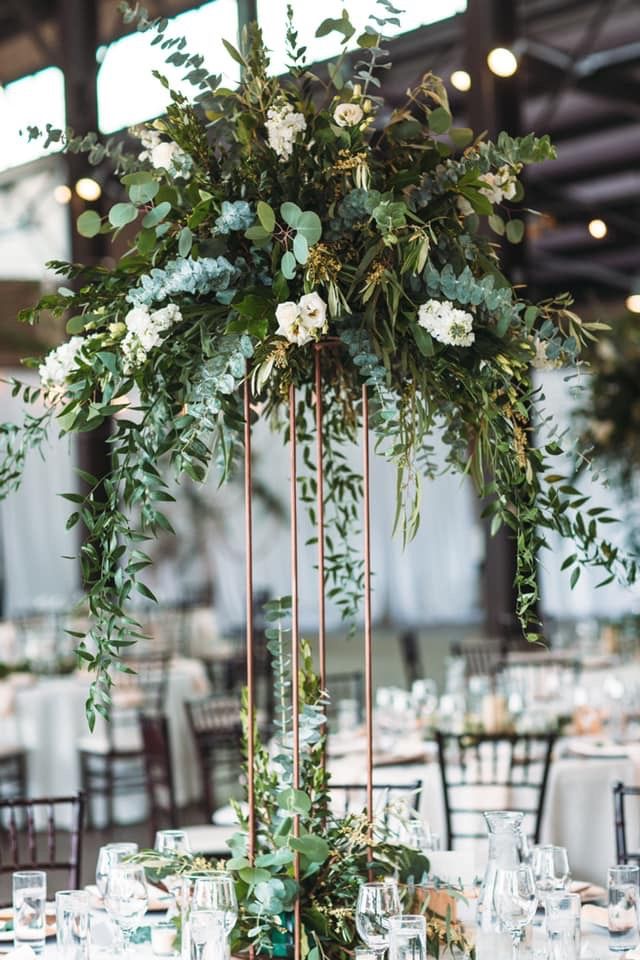 a tall centerpiece with greenery and white flowers is on top of a table