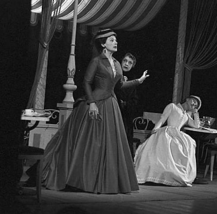 two women in long dresses are talking to one another on the stage with other people