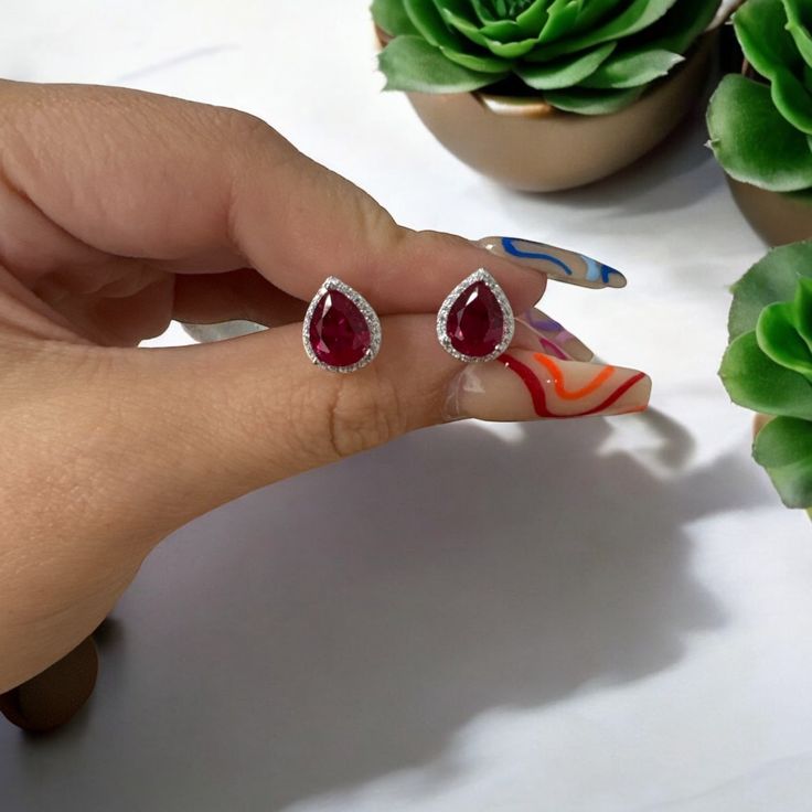 These Theodora Ruby Teardrop Gemstone Stud Earrings are a true marvel, each earring showcases a lab-created ruby that gleams with deep, vibrant hues, nestled within a teardrop silhouette.   Surrounding this central ruby is a delicate row of glittering zirconia, adding an extra touch of sparkle to your look.  The earrings are designed for ease and comfort, featuring a classic post and butterfly fastening. Handcrafted in 925 Sterling Silver dipped in 1Rhodium. Lab grown gemstone. Cubic Zirconia. Pear-shaped Ruby Gemstone Earrings, Pear-shaped Ruby Earrings, Anniversary Drop Ruby Earrings, Red Gemstone Accented Drop Earrings, Ruby Teardrop Earrings Fine Jewelry, Pear-shaped Ruby Earrings For Gift, Ruby Teardrop Jewelry With Matching Earrings, Teardrop Ruby Jewelry With Matching Earrings, Red Pear-shaped Cubic Zirconia Earrings