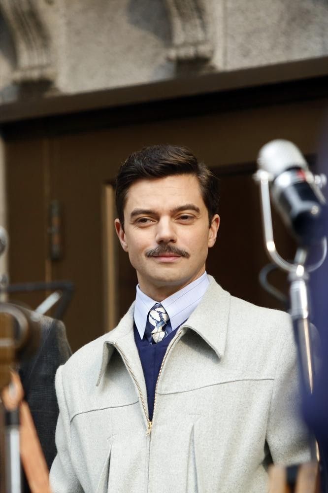 a man with a moustache standing in front of a microphone wearing a coat and tie