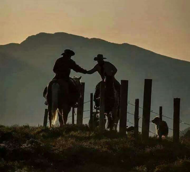 two men on horses standing next to each other near a fence and mountains in the background