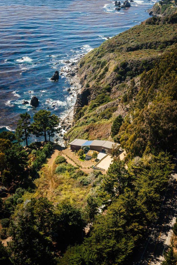 an aerial view of a house on the side of a cliff by the ocean with trees and rocks