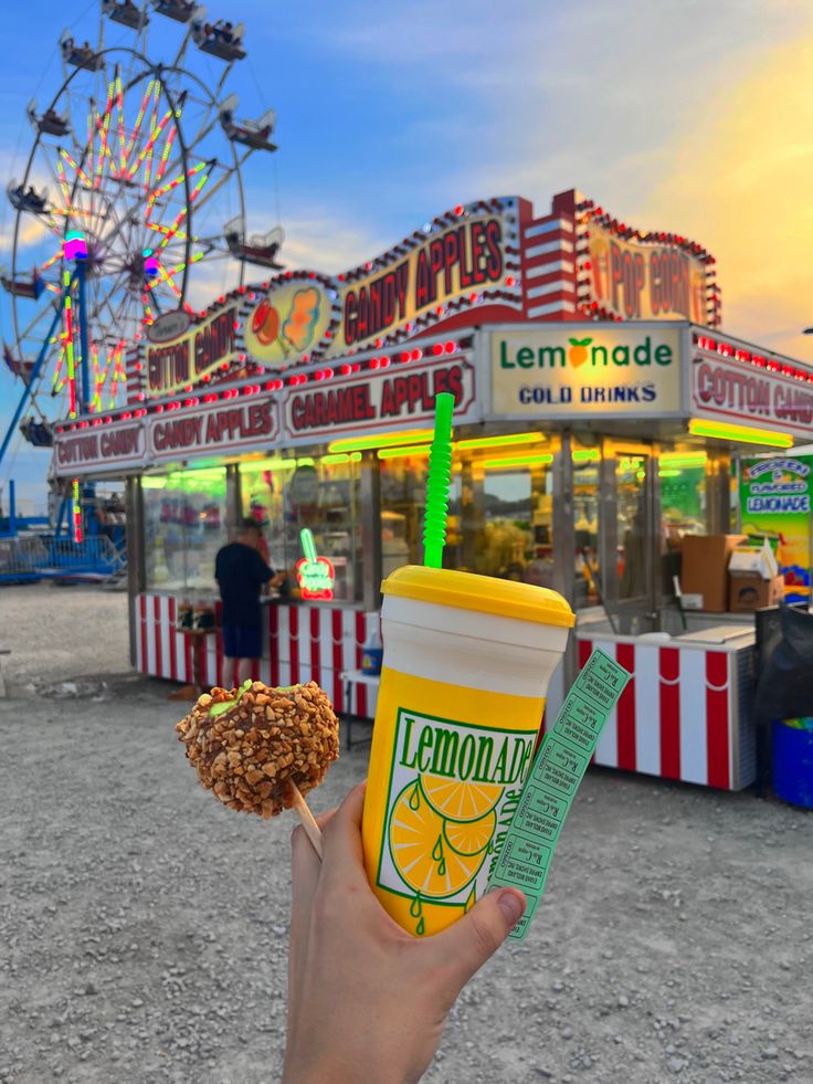 a person holding up a drink and donut in front of an amusement park ride