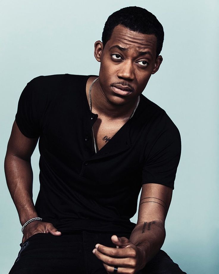 a man sitting down with his hands on his knees and looking at the camera while wearing a black shirt