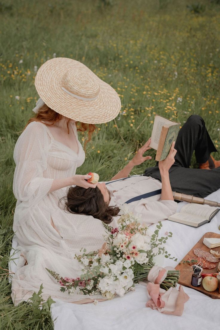 a woman in a white dress and straw hat sitting on a blanket reading a book