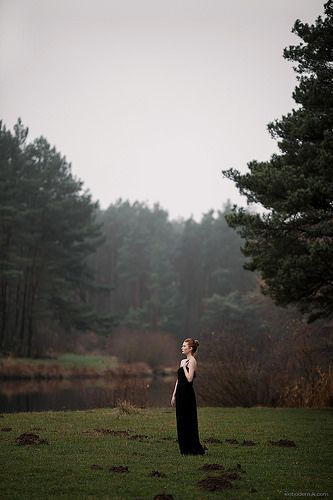 a woman in a long black dress is standing on the grass with trees behind her