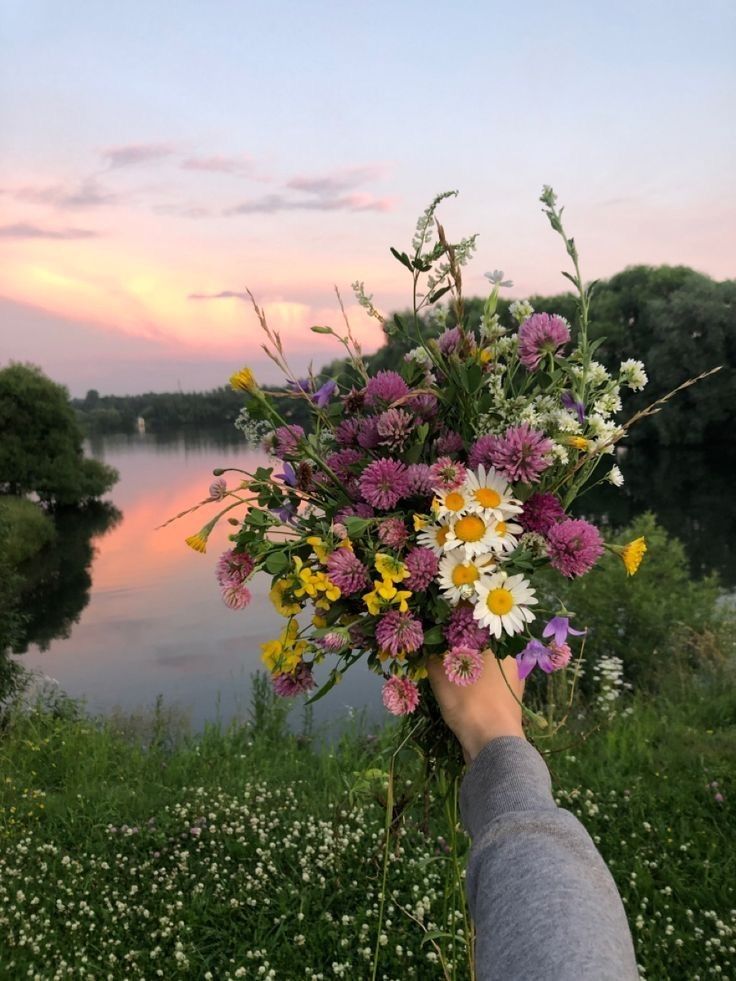 a person's hand holding a bouquet of wildflowers in front of a lake