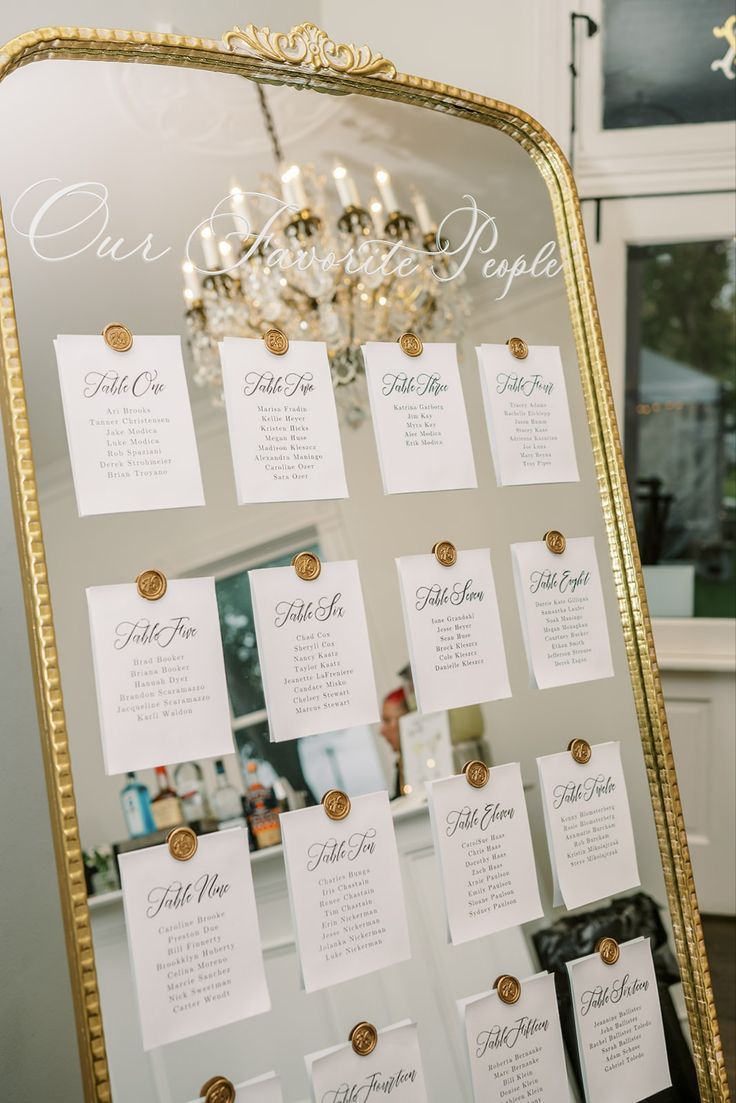 a gold framed mirror with seating cards attached to the front and back of it, along with a chandelier in the background