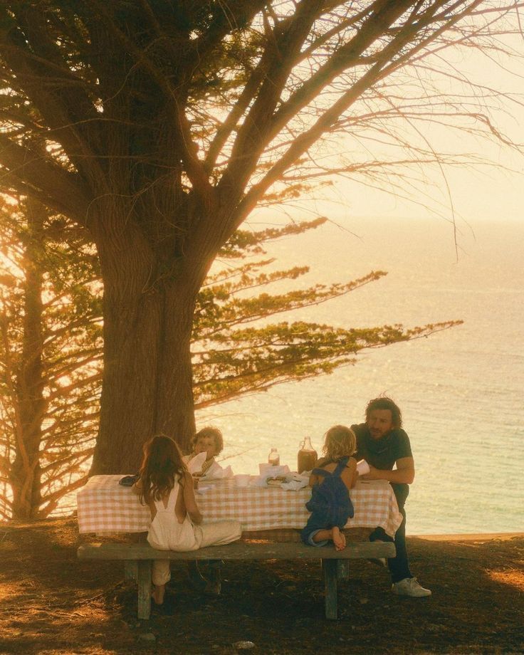 people are sitting at a picnic table by the water with their backs to each other