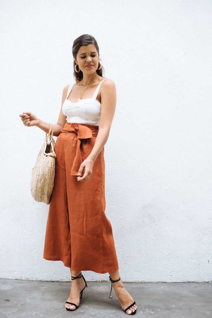 a woman standing in front of a white wall wearing an orange skirt and holding a straw bag