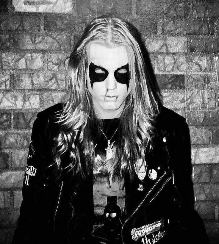 a man with long hair and black eye makeup wearing a leather jacket standing in front of a brick wall