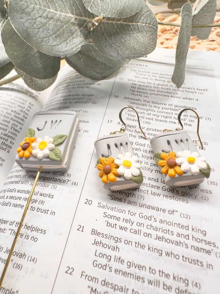 two small flower earrings sitting on top of an open book next to a green plant