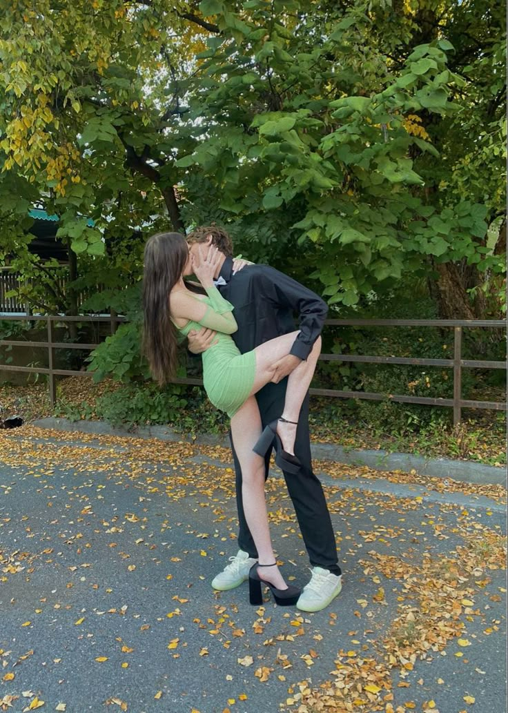 a man and woman kissing in front of trees with leaves on the ground behind them