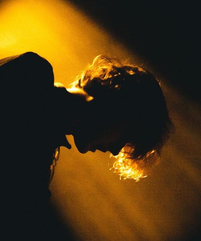 a man with long hair standing in front of a yellow light and holding his head to the side