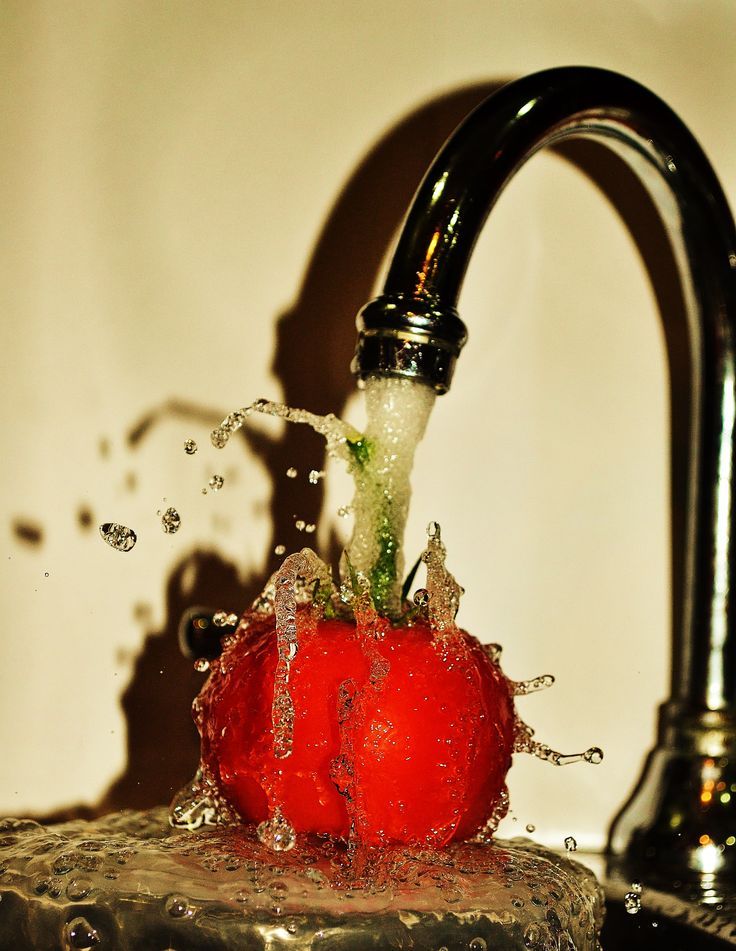 a red tomato being washed into a kitchen faucet with water splashing on it