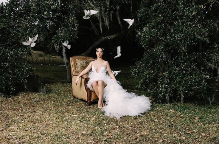 a woman in a white dress sitting on an old suitcase with birds flying around her