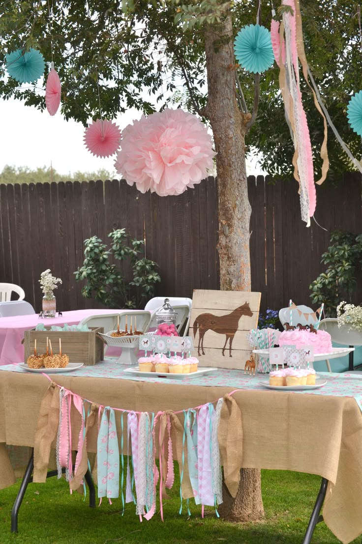 a table that has some cake on it in the grass with pink and blue tissue pom poms