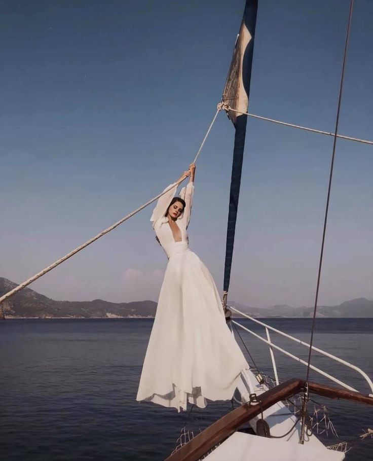 a woman in a white dress is standing on the front of a sailboat with her arms up