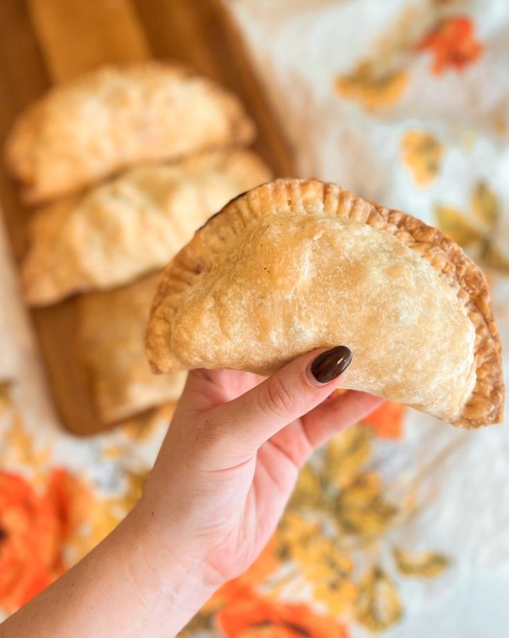 a hand holding a pastry in front of three other pastries on a floral tablecloth