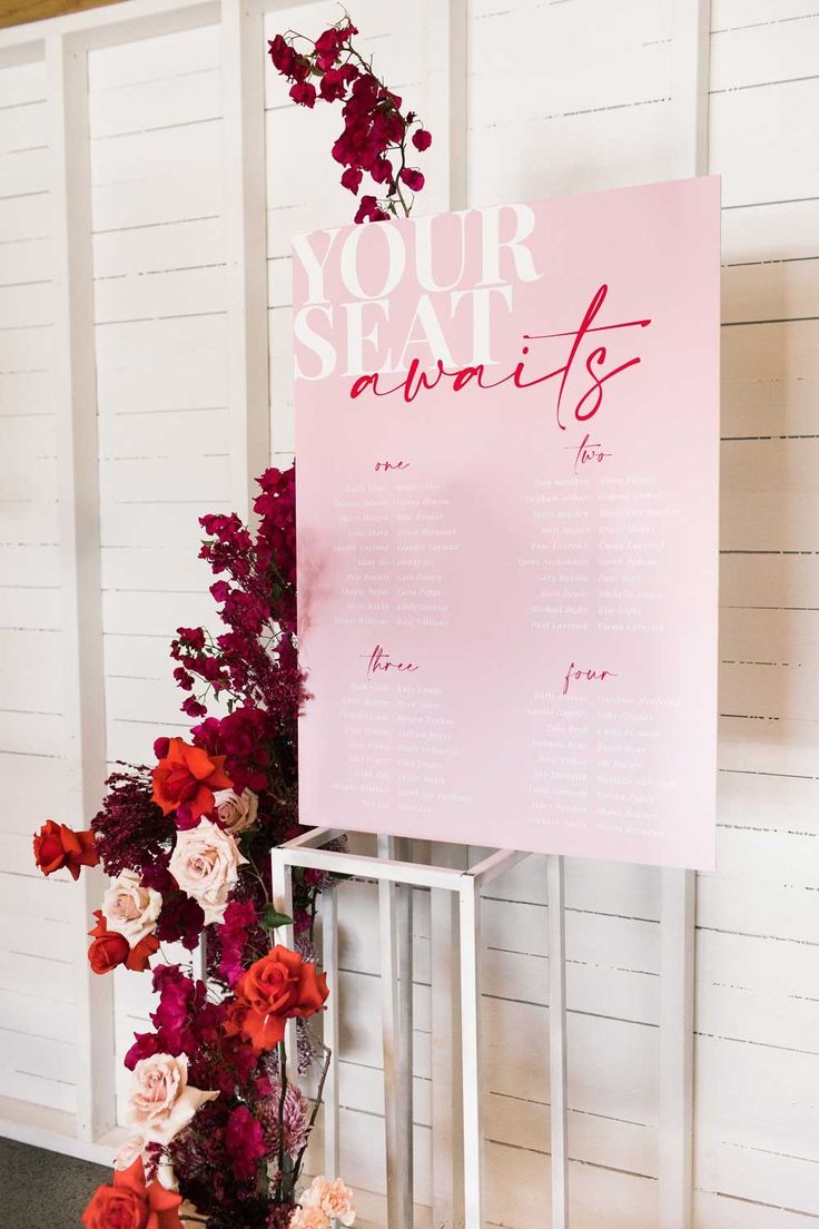 a sign that says your seat awaits with flowers in front of it on a stand