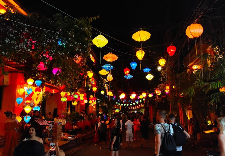 many people are walking down the street at night with colorful lanterns hanging from the ceiling