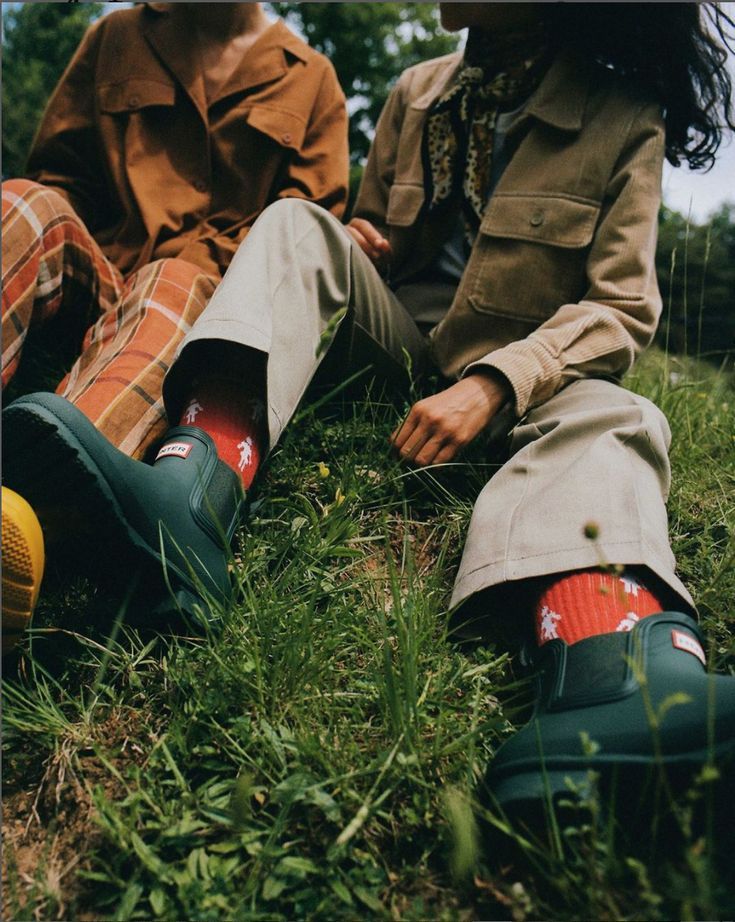 two people sitting in the grass with their feet up and one person wearing rain boots