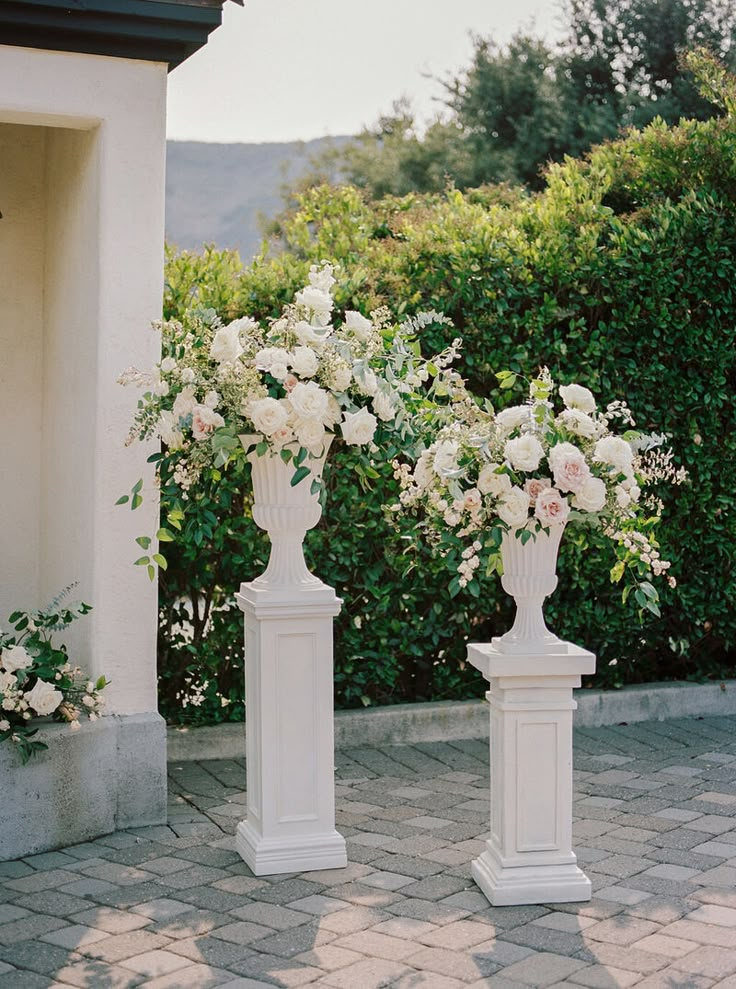 two white vases filled with flowers sitting on top of a brick floor next to bushes