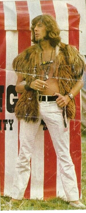 a man standing in front of a circus tent wearing white pants and fur vests