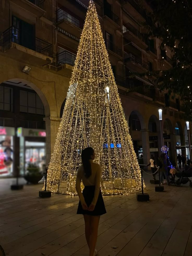 a woman standing in front of a lit up christmas tree on a city street at night